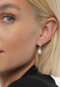 Náušnice s perlou a strieborným akcentom, obsahujúce hladkú, kvapkovito tvarovanú perlou a diamantový priehľadný kameň nad ňou.