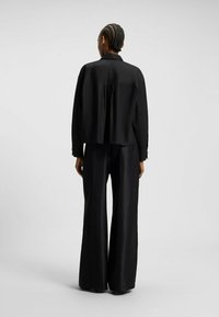 Donna con capelli intrecciati che indossa una maglietta nera a maniche lunghe larga e pantaloni neri a gamba larga, in piedi con la schiena rivolta verso uno sfondo neutro.