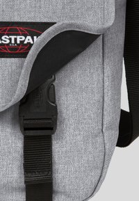 Grå tygbudväska med en texturerad yta, svart rem, spänne och röd "Eastpak"-logotyp på klaffen.