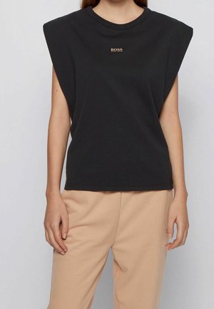Femme portant un haut sans manches noir à épaules rembourrées avec un petit logo doré "BOSS" et un pantalon beige, sur un fond uni.