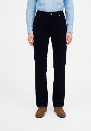 Personne portant un pantalon en velours côtelé noir, une chemise bleu clair à manches longues et des chaussures en daim marron, debout devant un fond blanc uni.