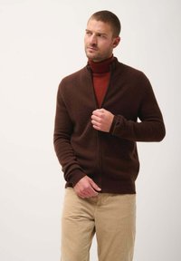 Cardigan marrone con zip realizzato in materiale morbido, con colletto rialzato e polsini a coste, indossato sopra un dolcevita rosso e pantaloni chiari.