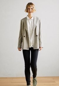 Blazer xadrez em cinza e branco, design de duplo botão, usado sobre uma camisa branca, combinado com jeans skinny pretos e sapatos verdes.