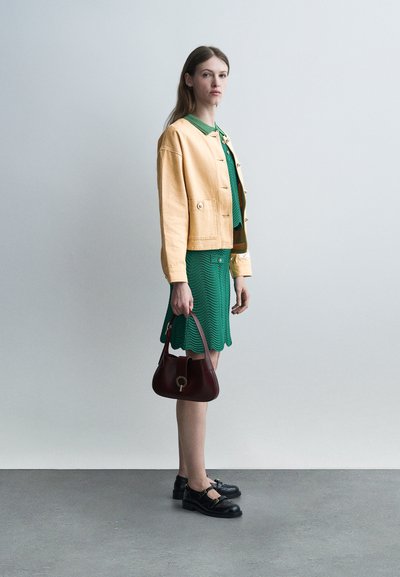 Νεαρή γυναίκα με πράσινο φόρεμα, μπεζ μπουφάν, μαύρα παπούτσια, κρατώντας καφέ τσάντα, στέκεται μπροστά σε απλό ανοιχτό γκρι φόντο.