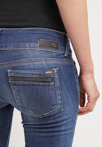 Blue denim jeans with a fitted design, featuring a brown leather tag, contrasting stitching, and a small zippered pocket on the back.
