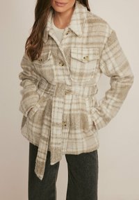 Beige- en wit geruit jasje met een ceintuur in de taille, gemaakt van zachte, gestructureerde stof, voorzien van twee borstzakken en grote knopen.