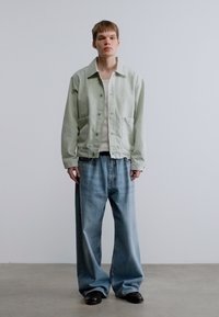 Φωτεινό πράσινο τζιν μπουφάν πάνω από λευκό πουκάμισο, σε συνδυασμό με άνετο, ανοιχτό μπλε τζιν. Η εμφάνιση χαρακτηρίζεται από χαλαρή εφαρμογή και διακριτή υφή.
