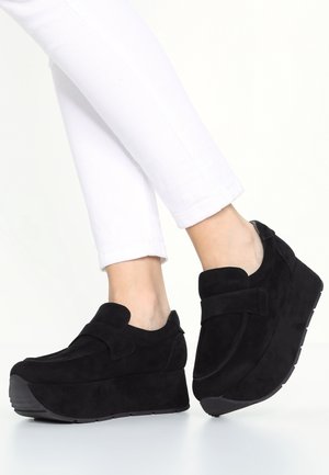 Osoba nosząca czarne buty na platformie typu slip-on w połączeniu z białymi dopasowanymi spodniami, pokazana od kolan w dół na białym tle.