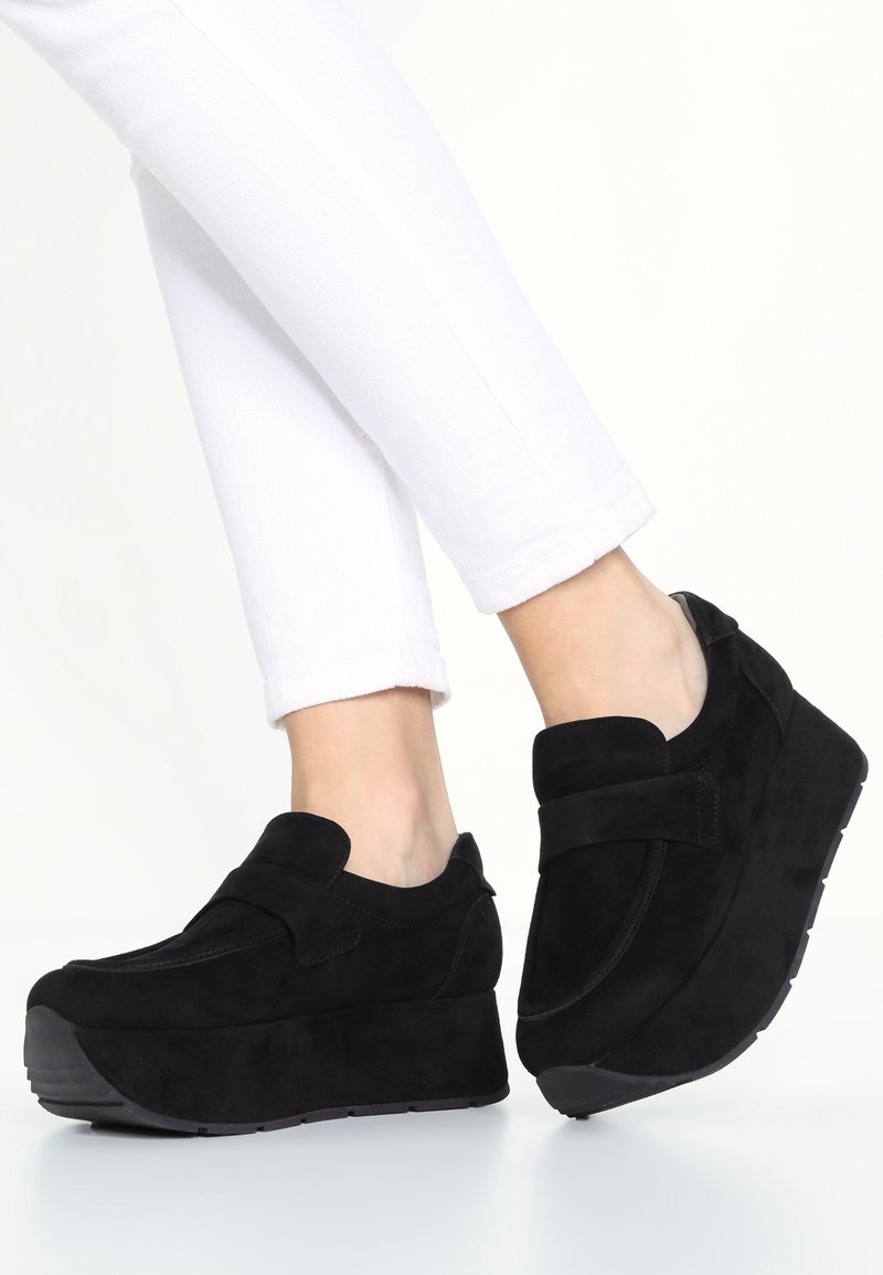 Personne portant des chaussures à plateforme noires sans lacets associées à un pantalon blanc ajusté, montrée des genoux vers le bas sur fond blanc.