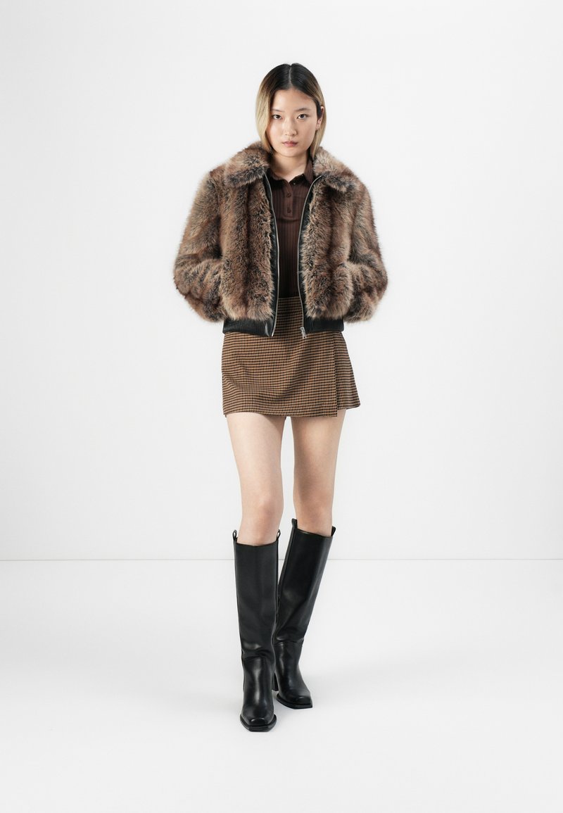 Kobieta w brązowej kurtce z sztucznego futra, brązowej mini spódnicy w wzory, ciemno-brązowej bluzce i długich czarnych skórzanych butach, stojąca na jednolitym tle.