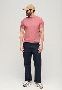 T-shirt rosa chiaro a maniche corte, pantaloni blu navy, cappellino beige, sneakers grigie con dettagli. In posa in piedi su uno sfondo neutro.