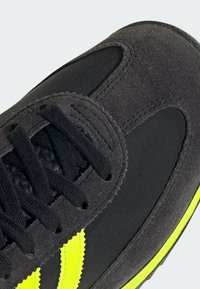 Černá sportovní obuv s kombinací svršku z semiše a nylonu. Obsahuje neonově žluté akcenty a černé tkaničky, což podtrhuje elegantní design a textura.