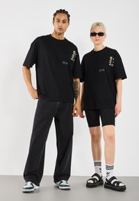 Des t-shirts noirs surdimensionnés avec des accents de logo, portés avec un jean foncé et des shorts de vélo noirs, complétés par des sandales et des chaussettes de sport.