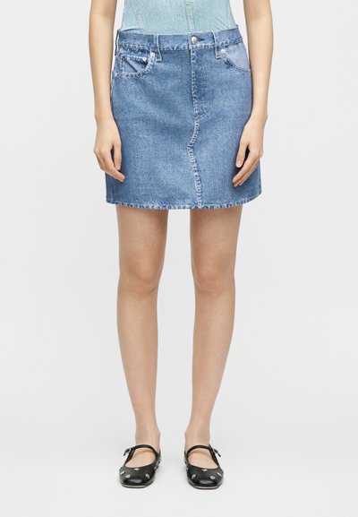 Γυναίκα που φοράει μια μπλε κοντή φούστα από τζιν με μπροστινές τσέπες και μαύρα παπούτσια Mary Jane με μεταλλικά τρουκς, στέκεται μπροστά σε απλό φόντο.