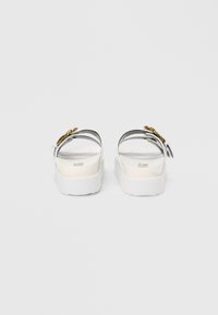 White sandals with a thick sole, transparent straps, and gold accents. Smooth texture with a slight sheen. Two adjustable ankle straps.