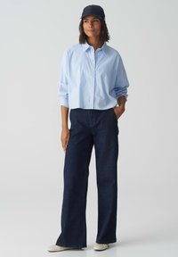 Camicia azzurra a strisce, con bottoni e maniche lunghe, abbinata a jeans a gamba larga blu scuro e un cappellino nero. Tessuto morbido con vestibilità casual.