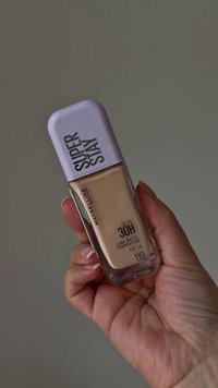 Super Stay flytande foundation i glasflaska, beige färg, med vit kork. Texten inkluderar "MAYBELLINE", "UP TO 30H" och SPF 16.