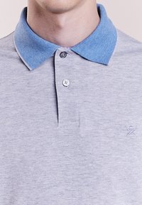 Polo shirt grigio chiaro con colletto in denim blu. Presenta due bottoni al collo e un logo ricamato sul petto. Tessuto morbido.