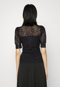 Schwarze Spitzenbluse mit kurzen Puffärmeln, taillierter Passform und gewelltem Saum. Der Rücken zeigt filigrane Blumenmuster.