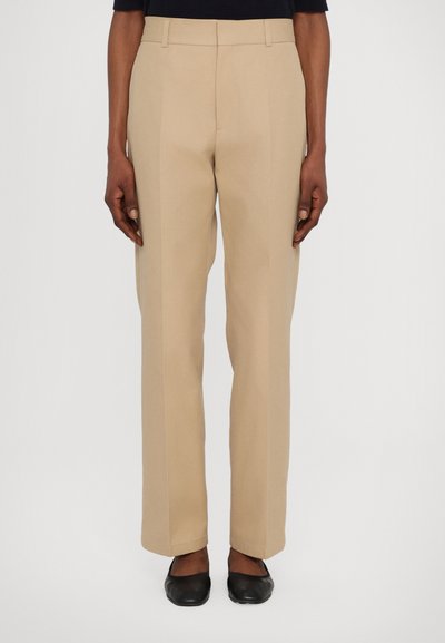 Beige maßgeschneiderte Hose mit geradem Bein, ausgestattet mit Gürtelschlaufen und Bügelfalten, kombiniert mit schwarzen flachen Schuhen.