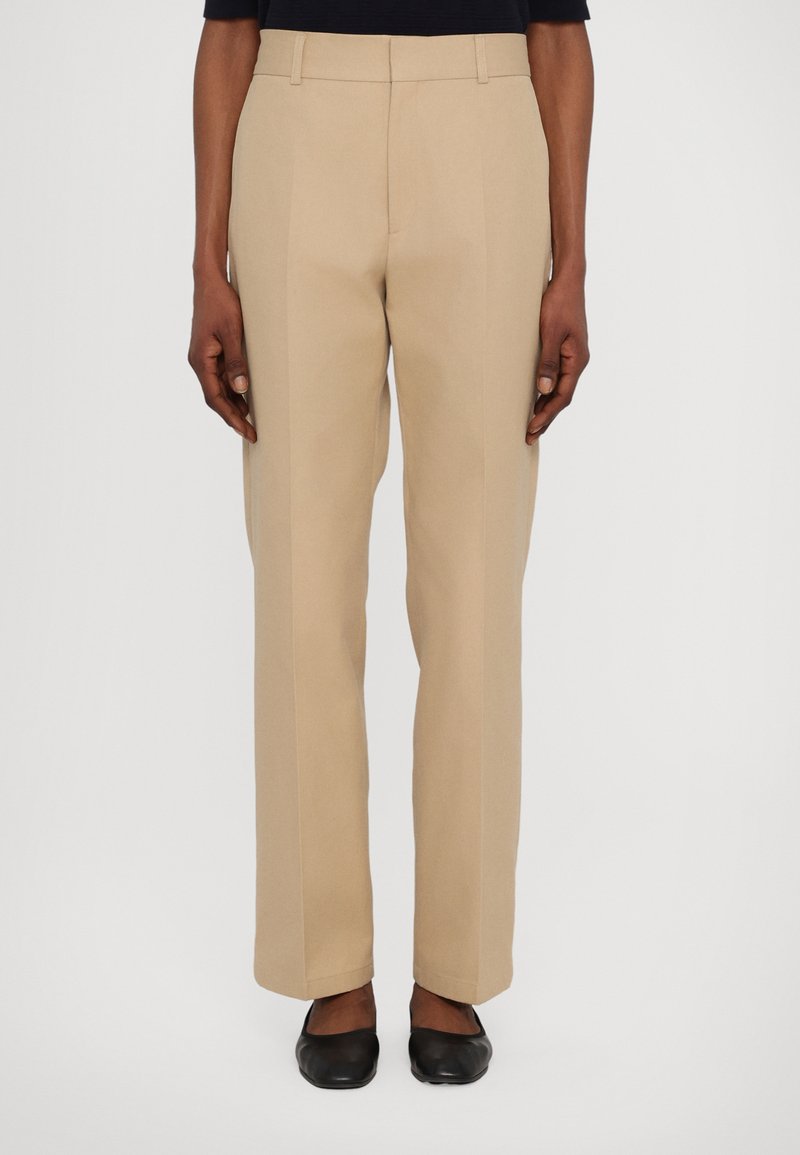 Pantaloni bej personalizați cu un design drept, având bucle pentru curea și detalii de plisare, asortați cu pantofi negri fără toc.