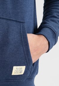Mano en el bolsillo delantero de una sudadera azul con una pequeña etiqueta rectangular beige que dice "BLEND" en la parte inferior delantera.