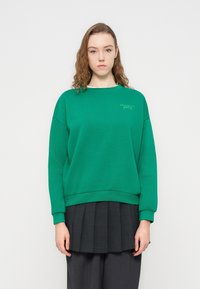 Πράσ ινο φούτερ με στρογγυλή λαιμόκοψη, ριμπ μανσέτες και τελείωμα. Διαθέτει διακριτική επωνυμία σε πιο ανοιχτή απόχρωση. Φοριέται πάνω από μια σκούρα πλισέ φούστα.