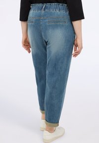 Pantalones de mezclilla de color azul claro con un corte relajado, cinturilla elástica, puños doblados y dos bolsillos traseros. El calzado visible incluye zapatos blancos.