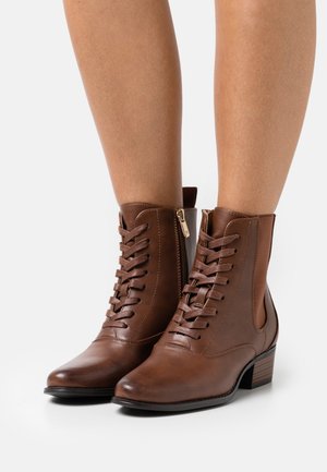 Bottines en cuir marron avec lacets, fermetures éclair latérales et petits talons empilés, portées sur des jambes nues sur fond blanc.