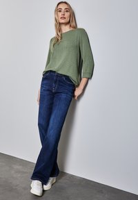 Zelený texturovaný svetr s tříčtvrtečními rukávy, spárovaný s tmavě modrými rozšířenými džíny a bílými teniskami, opřený o zeď.