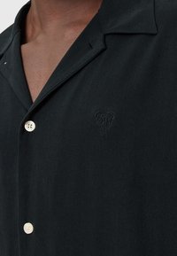 Chemise noire à manches courtes avec un col à revers, ornée d'un petit détail en cœur brodé et d'un seul bouton en perle. Texture de tissu lisse.