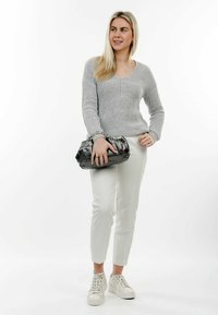 Maglione grigio a maglia con scollo a V, pantaloni bianchi su misura e sneakers bianche. Tiene in mano una borsa nera con texture e tracolla a catena.