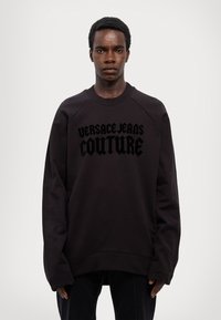 Crna dukserica od mekog materijala, s upečatljivim crnim grafičkim printom "VERSACE JEANS COUTURE" na prednjem dijelu, s raglan rukavima.