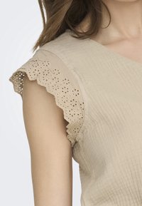 Top sans manches beige avec des bords en dentelle festonnée et des broderies florales en œillets sur l'épaule, porté par une personne avec de longs cheveux bruns.