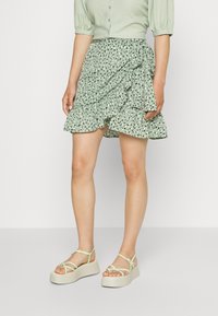 Jupe portefeuille verte à fleurs avec ourlet à volants, tissu léger, associée à des sandales compensées vert pâle avec fines brides et semelle texturée.