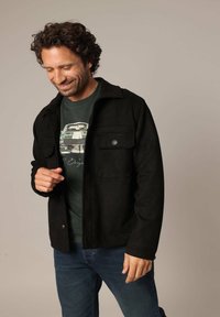 Veste en daim noire avec deux poches avant, fermeture à boutons et coupe décontractée, portée sur un t-shirt graphique vert et un jean bleu.