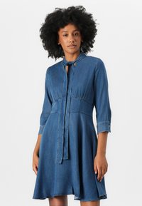 Denim dress with three-quarter sleeves, a fitted waist, and a flared skirt. Features a cut-out keyhole and tie detail at the collar.