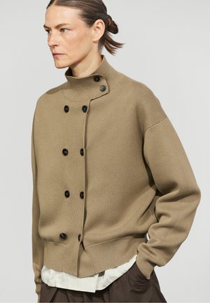 Donna con capelli raccolti che indossa un maglione beige con bottoni sopra una camicia bianca e pantaloni scuri, guardando leggermente a sinistra.