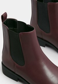 Bottines Chelsea en cuir bordeaux avec panneaux élastiques sur les côtés, semelles plates en caoutchouc noir, et texture lisse, dotées d'un tirant pour un enfilage facile.