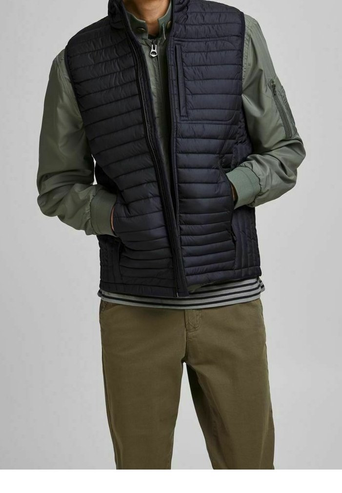 Homme portant un gilet matelassé noir par-dessus une veste zippée verte, les mains dans les poches du gilet, associé à un pantalon vert olive.