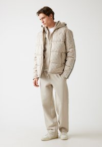 Jachetă puffern de culoare bej cu un model texturat, cu fermoar în față și glugă. Asortată cu pantaloni de trening de aceeași culoare și adidași albi, cu accente laterale.