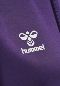Primo piano di un tessuto viola con un logo bianco raffigurante un'ape e la parola "hummel" sottostante, che enfatizza la texture liscia e il forte contrasto.