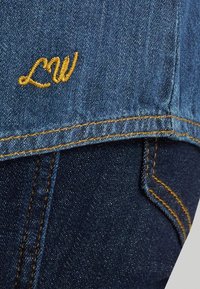 Ύφασμα τζιν με κίτρινα κεντημένα αρχικά "LW" πάνω από έντονη ραφή αντίθετου χρώματος, δείχνοντας κοντινή λήψη της επικάλυψης μπουφάν και τζιν παντελονιού.