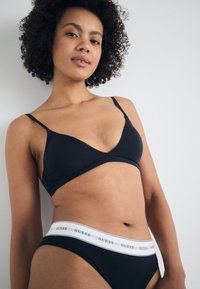Fekete bikini szett, amely sima, minimalista braletttel rendelkezik vékony pántokkal, és egy fehér GUESS logóval ellátott bikini alsóval.