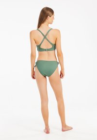 Zielony bikini z prążkowanego materiału, z krzyżowym zapięciem na plecach w górnej części oraz wiązanymi bokami w dolnej części, prezentujący gładką fakturę tkaniny.