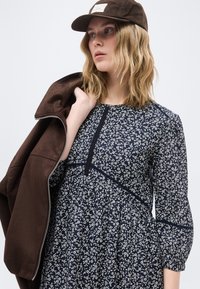 Νεαρή γυναίκα που φοράει καφέ καπέλο και μπλε φόρεμα με λουλούδια, κρατώντας ένα καφέ σακάκι στον ώμο της, κοιτάζοντας προς τα πλάγια.