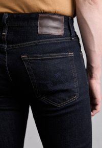 A sötét farmernadrág hátulján egy barna bőr folt található, a kontrasztos sárga varrás és a zseben lévő jellegzetes kék hímzés kiemelkedik.