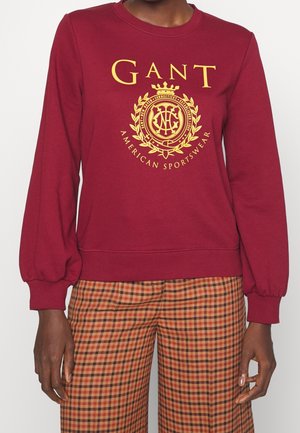 Person iført en vinrød sweatshirt med gul GANT-logo og emblem, kombineret med orange og sort ternede bukser.
