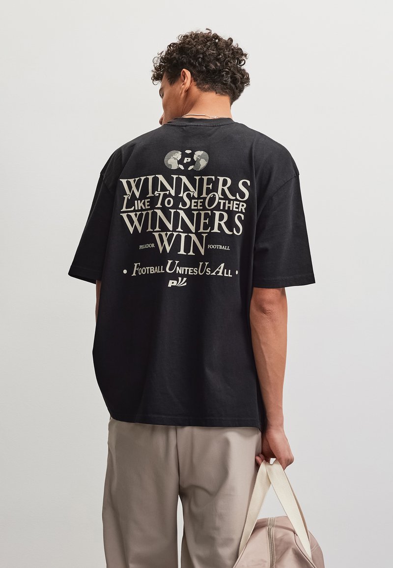 Άνδρας με μαύρο πουκάμισο με το κείμενο "Winners like to see other winners win" και κρατώντας μπεζ τσάντα σε απλό φόντο.