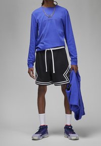 Camisa de manga larga azul, pantalones cortos de malla negros con ribete blanco, zapatillas moradas con detalles y sosteniendo una chaqueta azul.